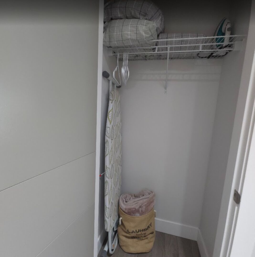 A laundry bag labeled 'LAUNDRY' in a corner of a room with a metal shelf above holding a pillow and an iron.