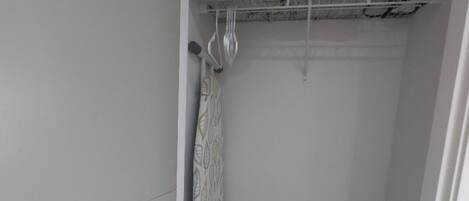 A laundry bag labeled 'LAUNDRY' in a corner of a room with a metal shelf above holding a pillow and an iron.