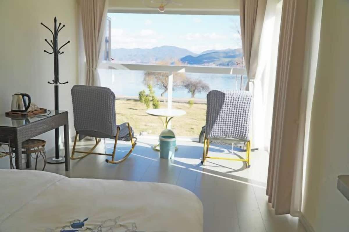 A bedroom with a bed, a coat rack, two rocking chairs, a small table with a kettle, and a view of a lake and mountains.