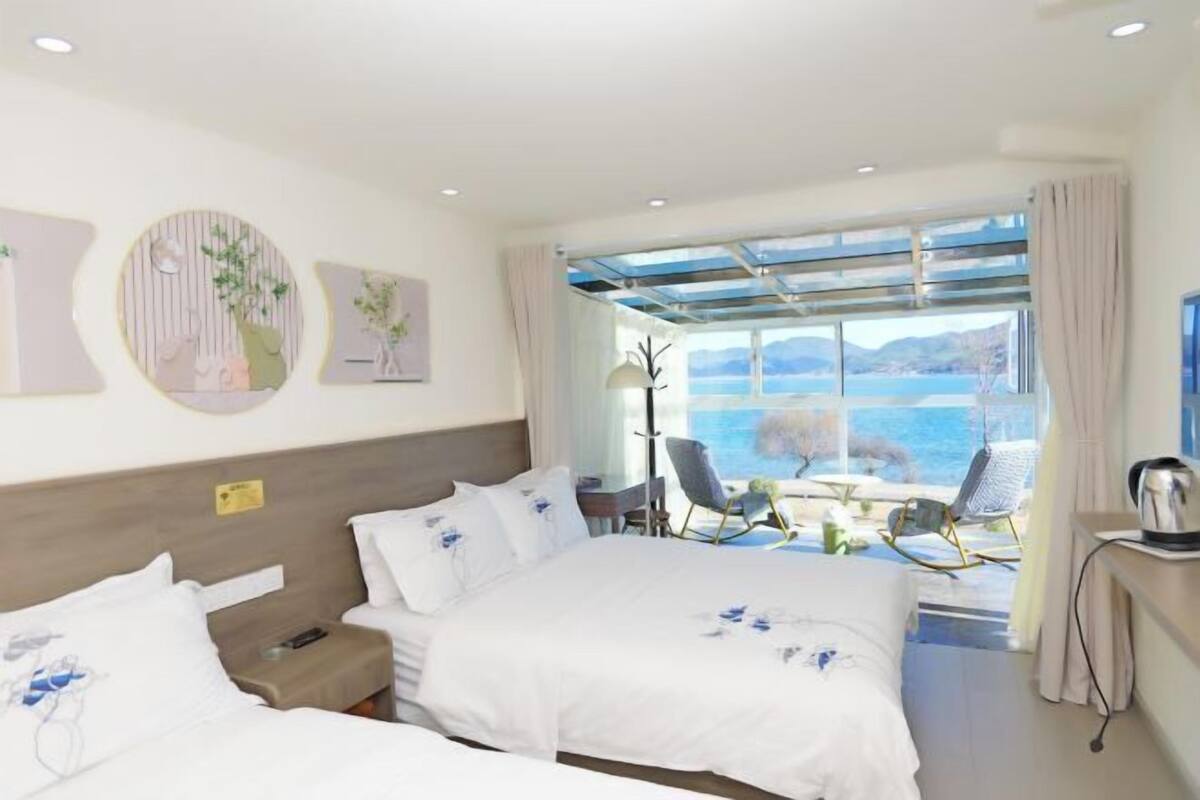 A hotel room with two beds, a desk, a chair, and a view of the sea.