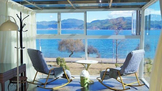 Une pièce avec un mur en verre, deux fauteuils à bascule, une petite table et une vue sur un lac et des montagnes.
