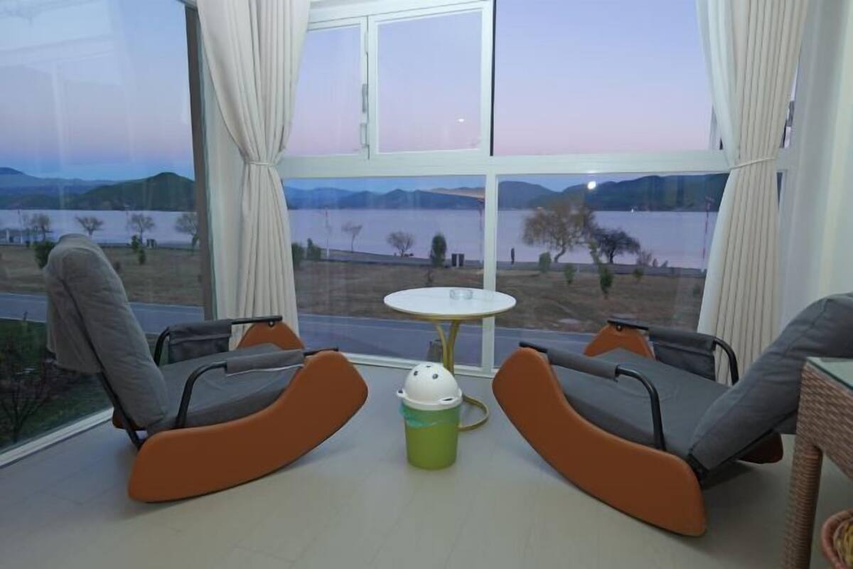 A room with two orange chairs, a small table, and a view of a lake and mountains through large windows.