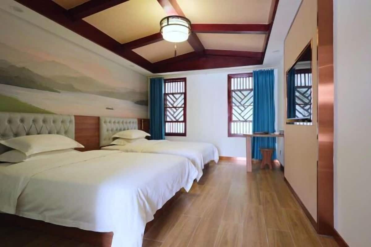 A hotel room with two beds, a wooden ceiling, and a view of the outdoors through a window with blue curtains.