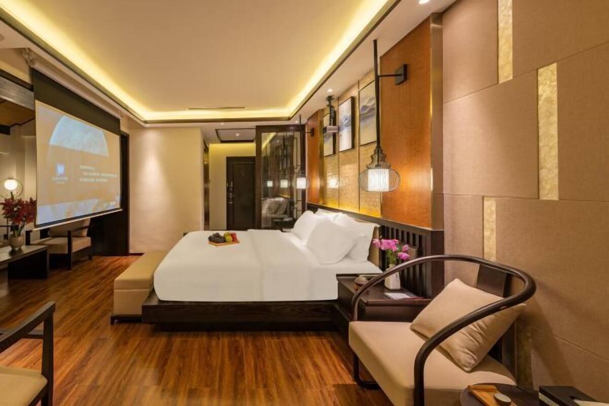 A modern hotel room with a large bed, a seating area with a sofa and armchair, a TV, and a wall-mounted lamp.