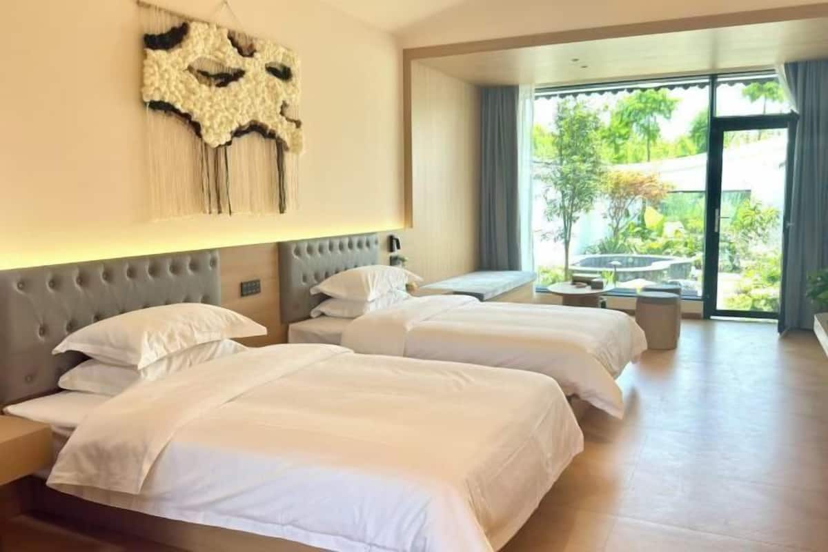 A hotel room with two beds, a large window with a view of greenery, and a seating area with a table and chairs.