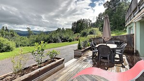 Una terraza de madera con mobiliario exterior, una silla roja y vistas a un paisaje verde con árboles y colinas.