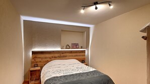 Un dormitorio con cabecera de madera, cama con ropa de cama estampada, mesita de noche, una pequeña mesa y estante con adornos.