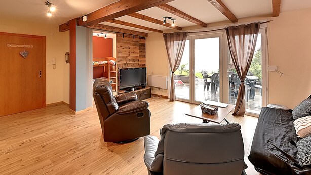 Una sala de estar con techo de madera, un sofá de cuero, un sillón reclinable, un televisor sobre un mueble bajo y un ventanal corredizo que da a un área exterior.