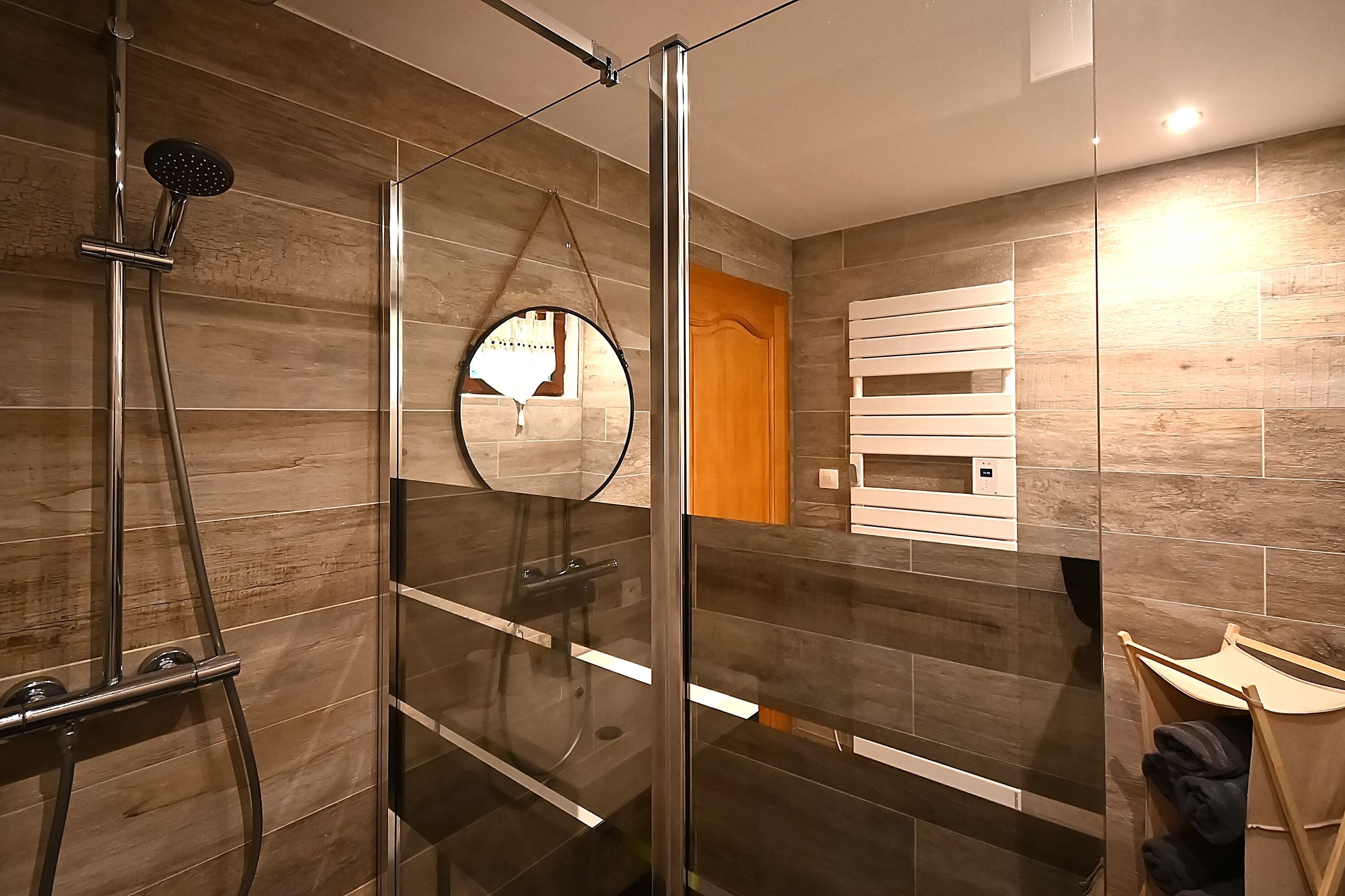 A modern bathroom with a glass-enclosed shower, a round mirror, and wooden wall tiles.