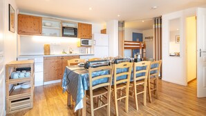 A compact kitchen with wooden cabinets, a microwave, and a refrigerator. There is a dining table with chairs and a small kitchen island with storage. - Le Machu - Appt 3*, vue montagne (Chamrousse)