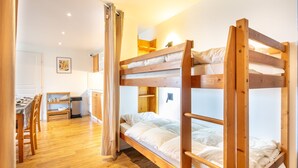 A bunk bed room with a wooden floor, a desk with chairs, and a small kitchen area. - Le Machu - Appt 3*, vue montagne (Chamrousse)