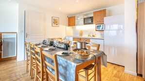 A dining area with a wooden table set for four, surrounded by chairs. A microwave, oven, and refrigerator are visible in the kitchen area. - Le Machu - Appt 3*, vue montagne (Chamrousse)