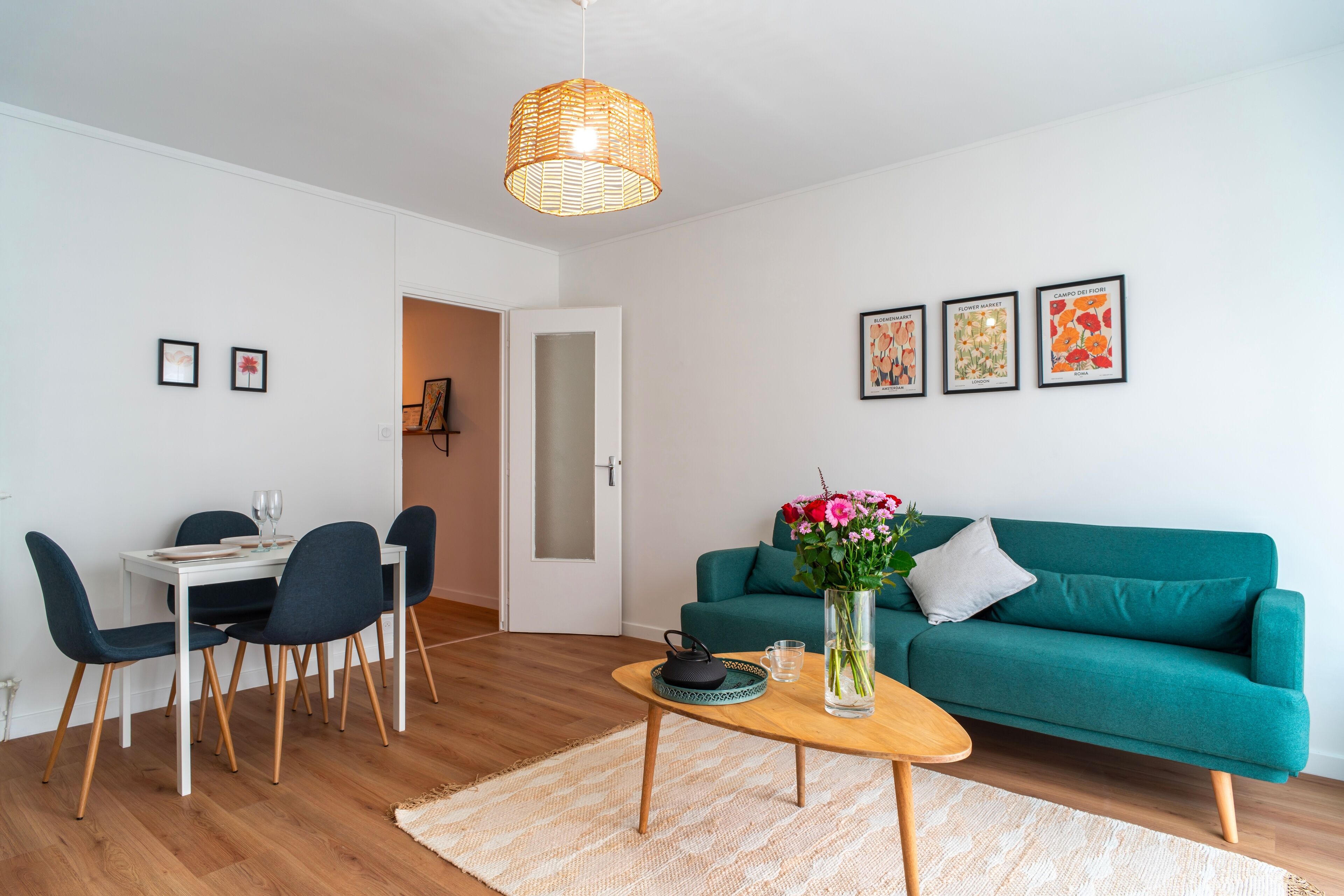 A modern living room with a teal sofa, a dining table with chairs, a coffee table with a vase of flowers, and a hanging light fixture.