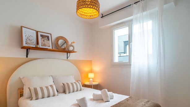 A neatly made bed with pillows, a bedside lamp, and a window with sheer curtains.