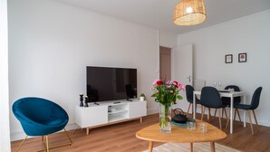 A modern living room with a flat-screen TV, a blue chair, a wooden dining table, and a vase of flowers.