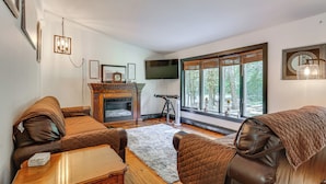 A cozy living room with leather sofas, a fireplace, a flat-screen TV, and large windows with a view of trees.