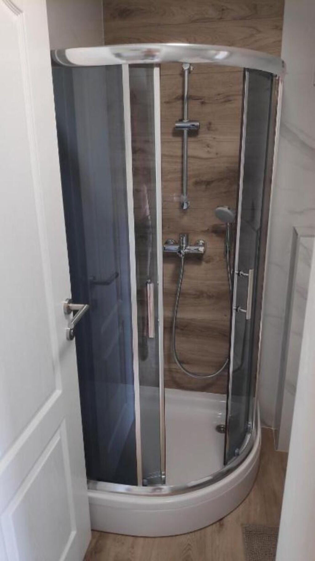 A glass-enclosed shower stall with a wooden floor, a showerhead, and a handheld shower attachment.