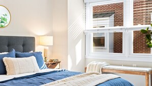 Une chambre à coucher avec un lit, une table de chevet avec une lampe, une fenêtre donnant sur un immeuble en briques et une plante posée sur le rebord de la fenêtre.