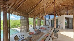Uma ampla área de estar ao ar livre com telhado de palha, piscina e um espaço de convívio com sofás e cadeiras.