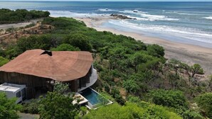Um imóvel à beira-mar com uma grande estrutura de telhado de palha, piscina e vegetação exuberante.