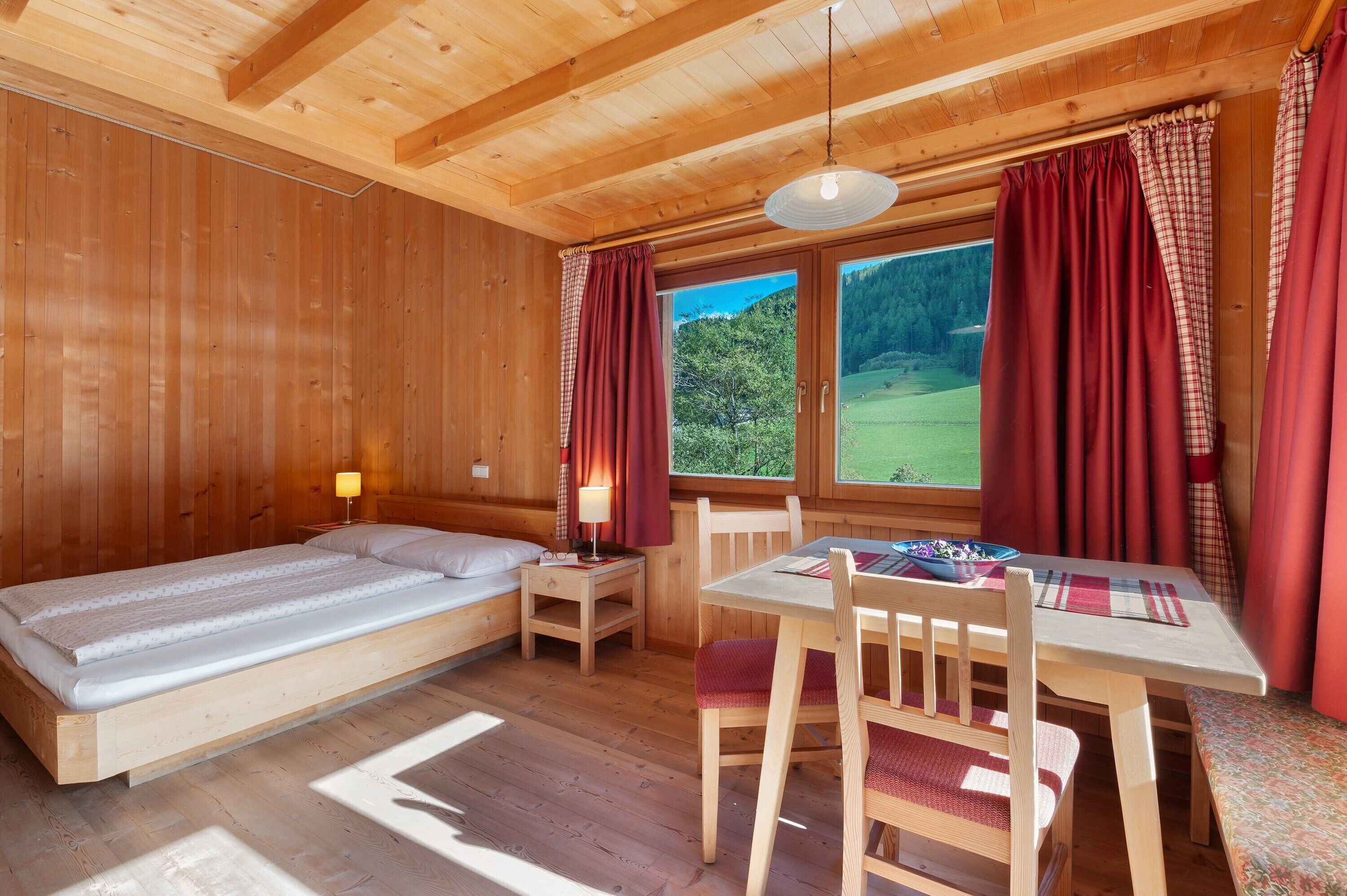 A wooden cabin with a bed, a table with chairs, a lamp, and a view of the outdoors.