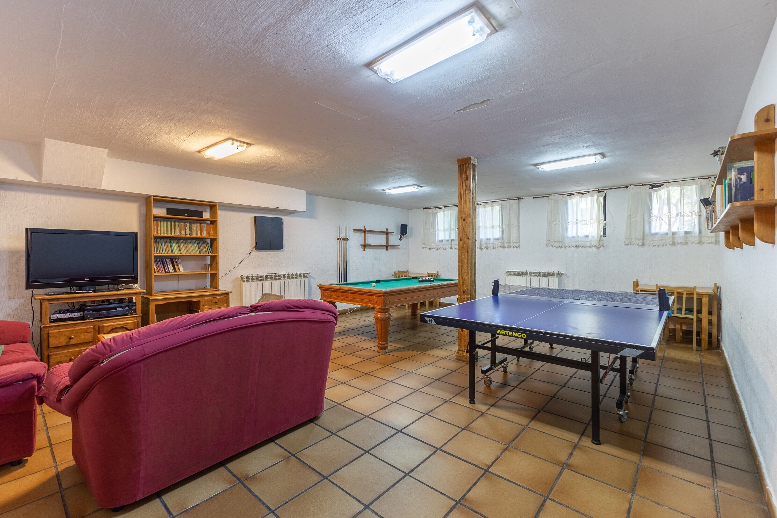 Um quarto com mesa de bilhar, mesa de tênis de mesa, sofá, televisão e estante de livros.