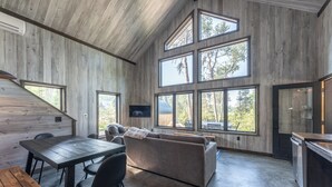 En rummelig opholdsstue med træloft, store vinduer, sofa, spisebord og et køkkenområde.