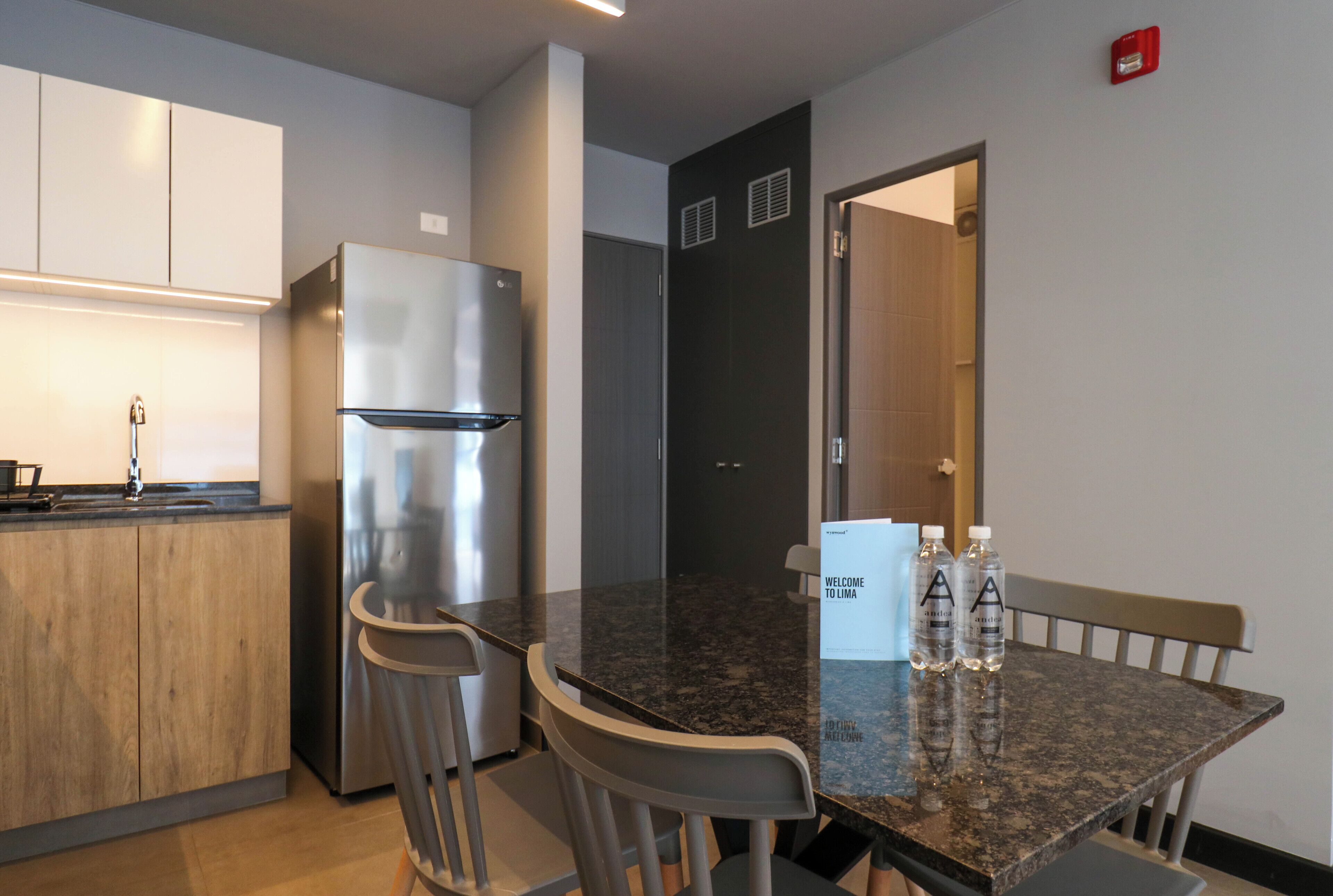 A modern kitchen with a stainless steel refrigerator, wooden cabinets, and a granite countertop. There are two bar stools and a welcome sign.