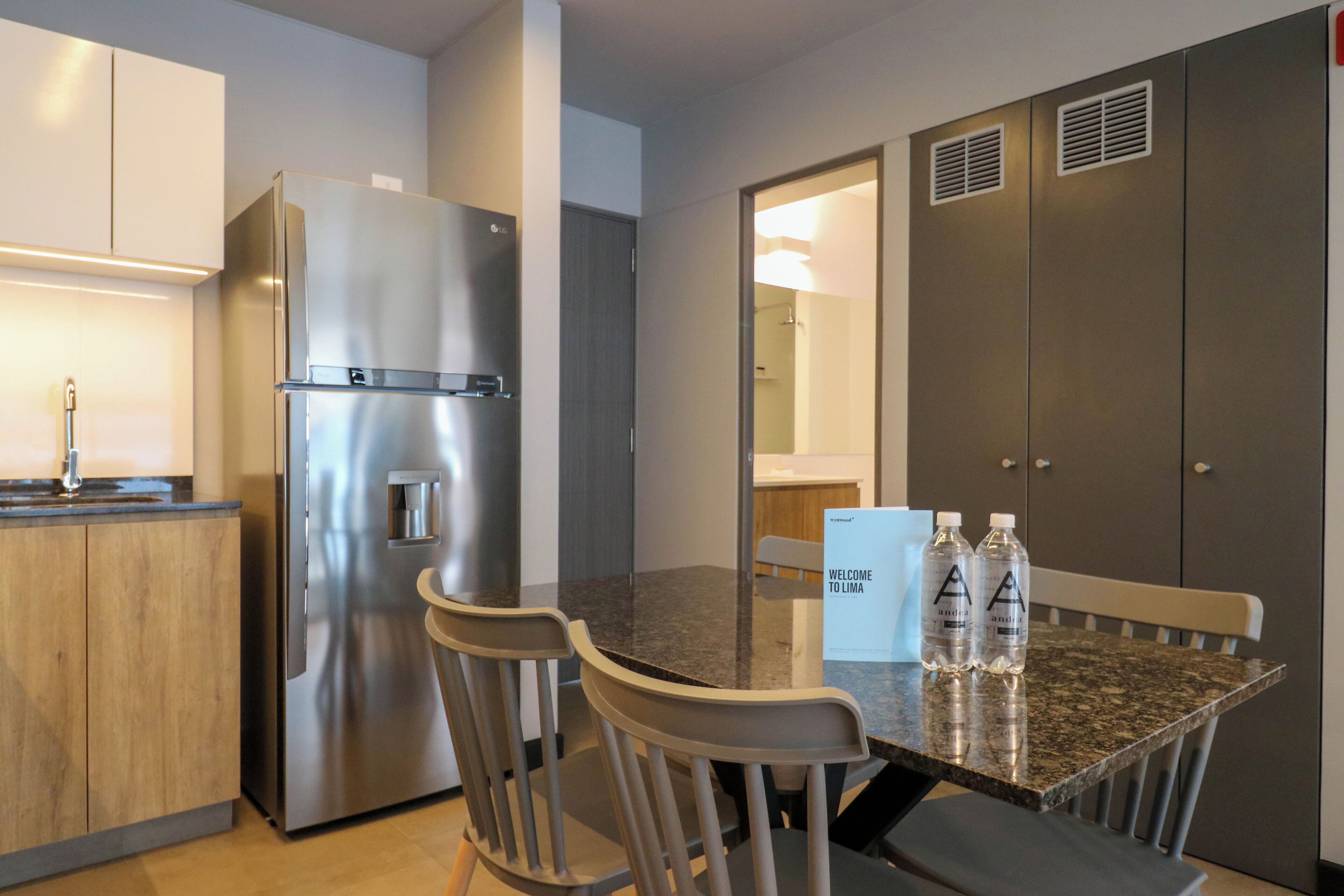 A modern kitchen with a stainless steel refrigerator, wooden cabinets, a granite countertop, and a dining area with two chairs and a table.