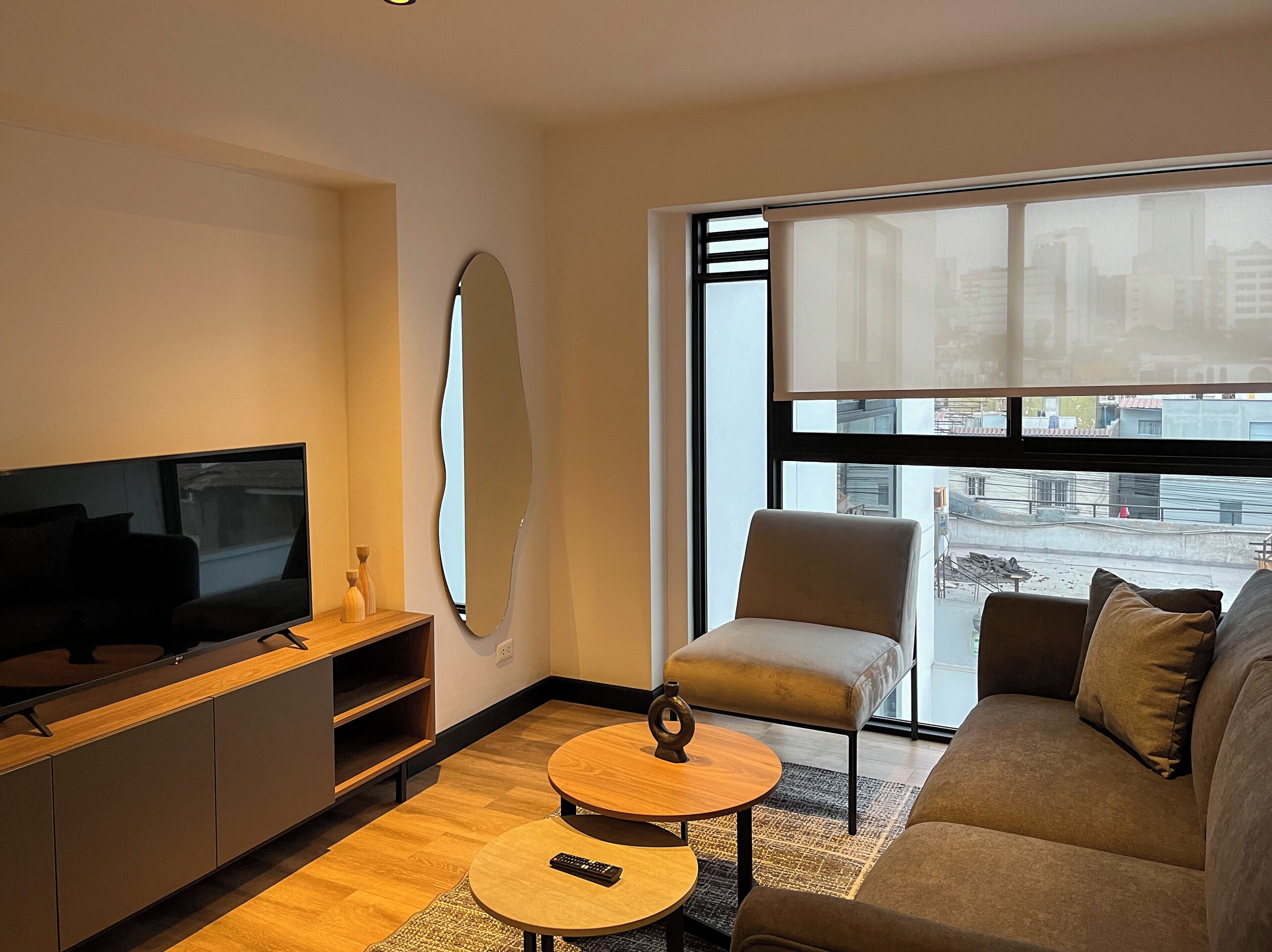 Ein modernes Wohnzimmer mit Flachbildfernseher, rundem Couchtisch, Sofa, Sessel und einem großen Fenster mit Blick auf Gebäude.