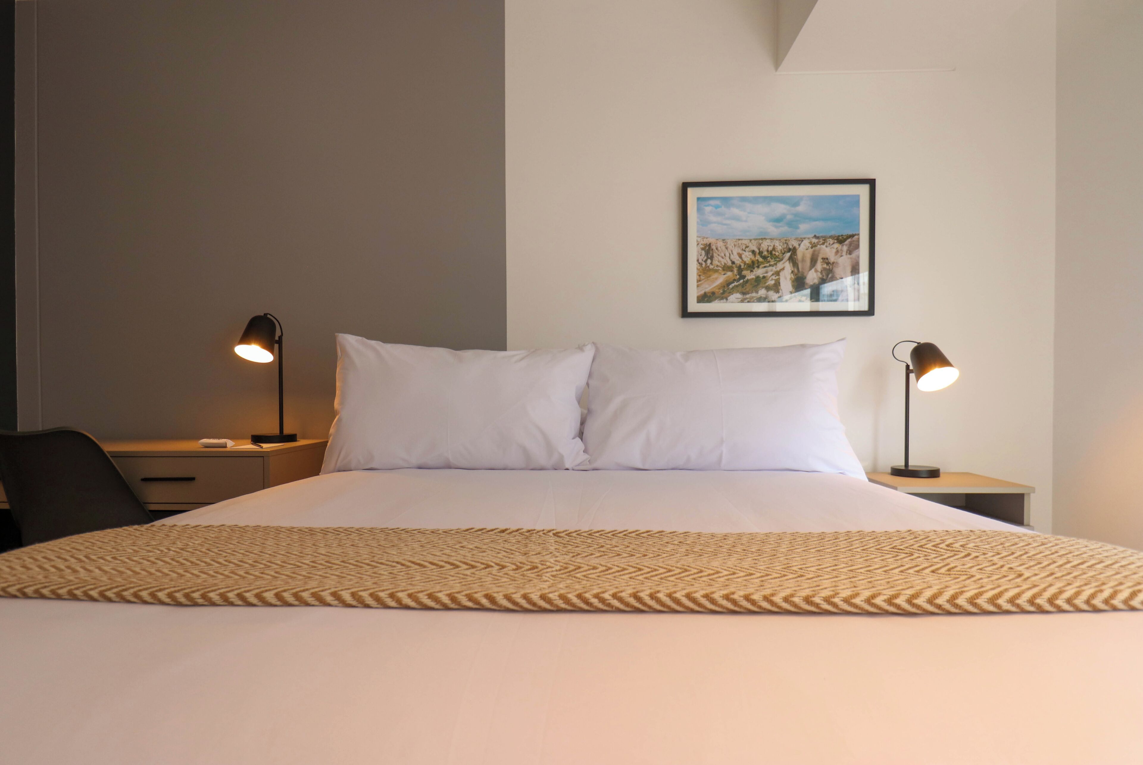 Una cama bien hecha con sábanas blancas y un plaid estampado, flanqueada por dos mesitas de noche con lámparas, en una habitación con un cuadro en la pared.