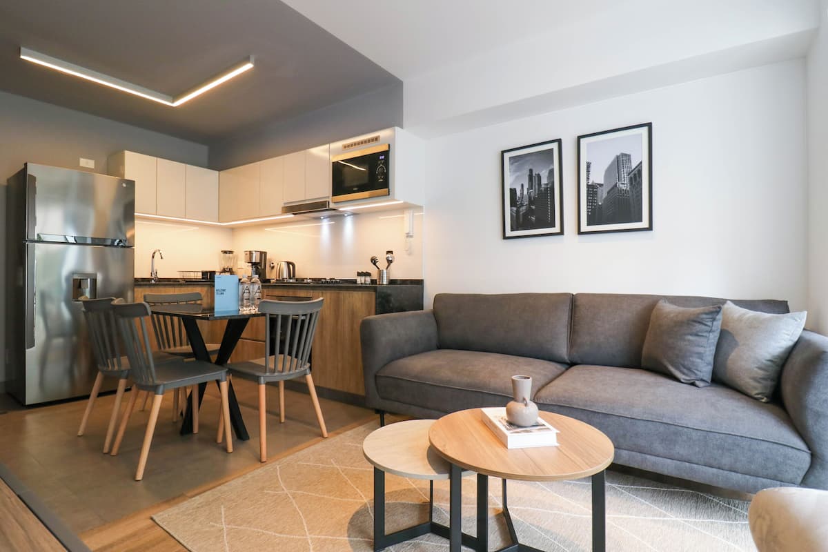 Eine moderne Küche mit Kühlschrank, Mikrowelle und Essbereich. Ein graues Sofa und ein kleiner Tisch mit einer Vase stehen dort. Zwei gerahmte Bilder hängen an der Wand.