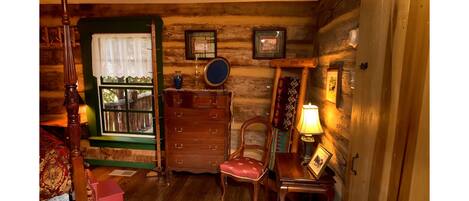 A cozy log cabin bedroom with a wooden bed, a dresser, a chair, and a small table with a lamp.