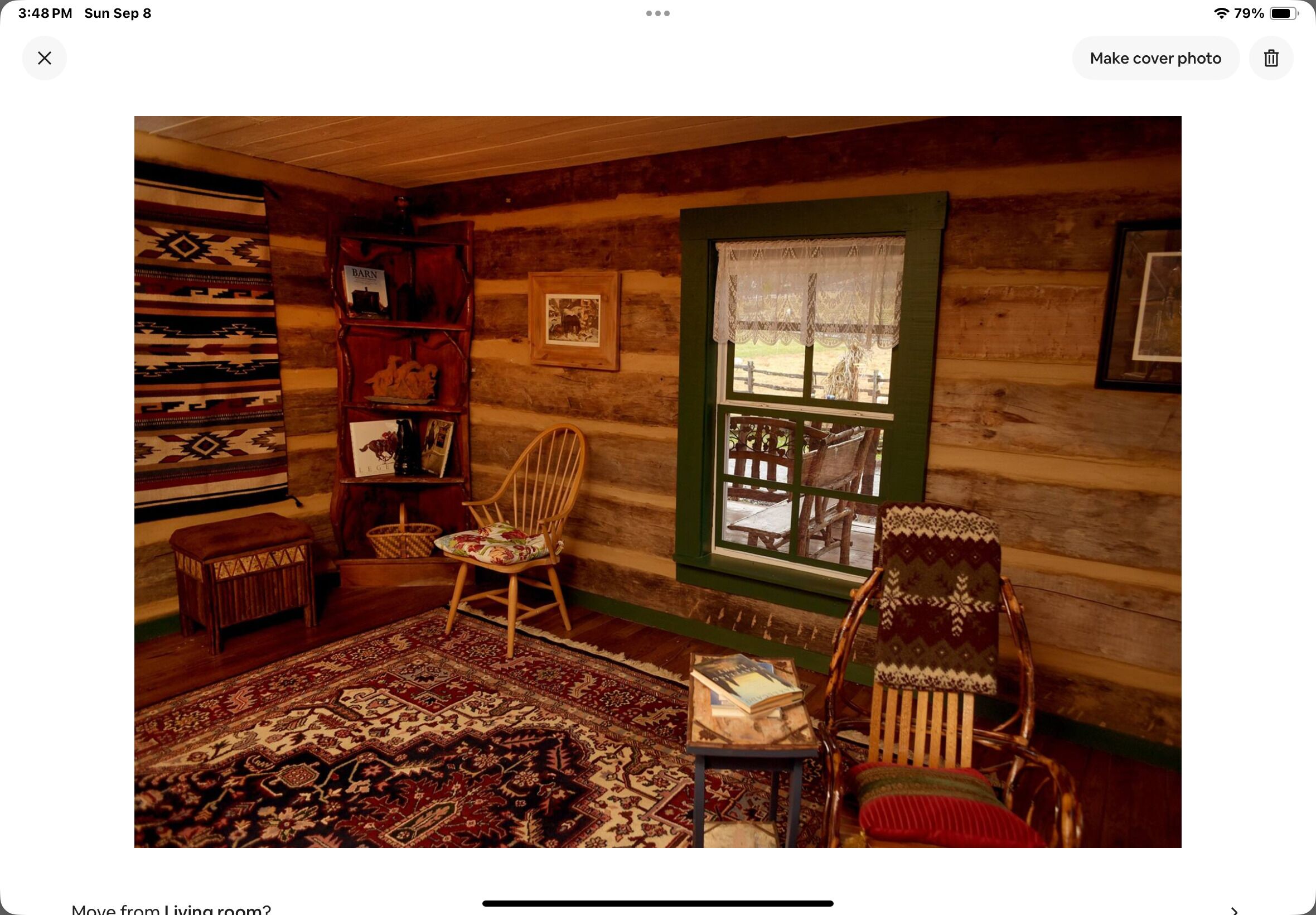 A cozy log cabin interior with a wooden bookshelf, two chairs, a patterned rug, and a window with a view of the outside.