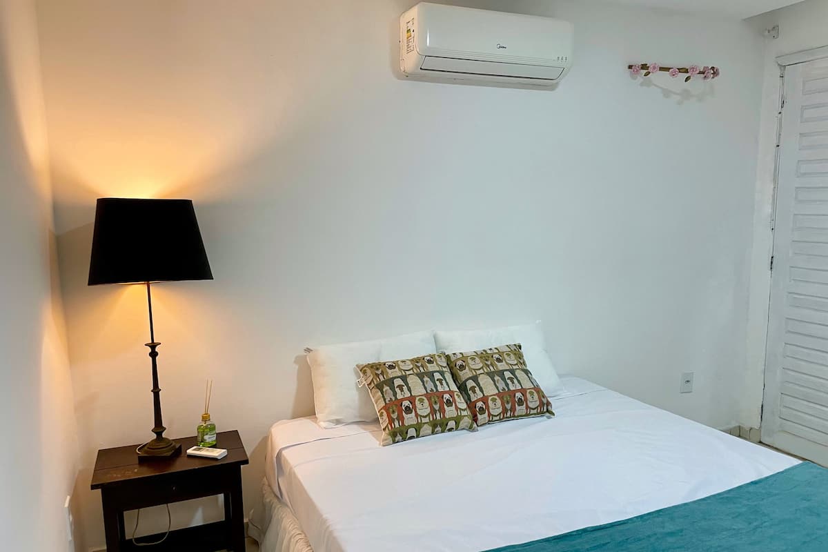Un dormitorio con una cama, una mesita de noche con lámpara, un aire acondicionado instalado en la pared y una puerta blanca.