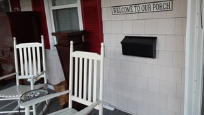 Une véranda avec deux fauteuils à bascule blancs, une boîte aux lettres et une pancarte indiquant : « Bienvenue sur notre véranda ».