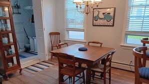 Un coin repas avec une table et des chaises en bois, un lustre, une fenêtre avec des stores et un tableau représentant un scooter accroché au mur.