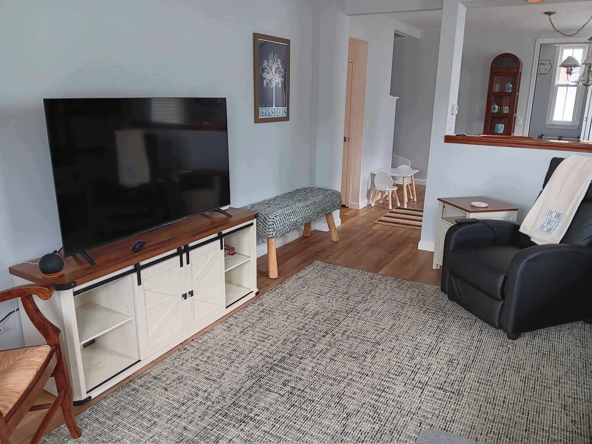 A living room with a flat-screen TV on a wooden stand, a bench, a black leather sofa, a patterned rug, and a dining area with a table and chairs.