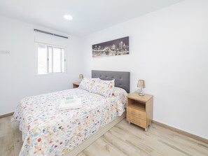 Quarto com cama, mesa de cabeceira, abajur, janela e pintura de paisagem urbana na parede.
