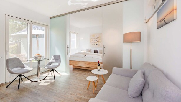 A modern hotel room with a glass door leading to a balcony, a bed with a wooden headboard, a grey sofa, and a small round table with a bottle and glass. - "Kuschelnest" Vacation Home: Modernly Furnished with a Sun Terrace - Cozy (Kamminke)