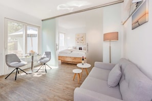 Une chambre d’hôtel moderne dotée d’une porte vitrée donnant sur un balcon, d’un lit avec une tête de lit en bois, d’un canapé gris et d’une petite table ronde sur laquelle se trouvent une bouteille et un verre.