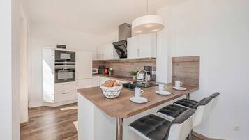 A modern kitchen with white cabinets, a wooden countertop, a built-in oven, a coffee maker, and a seating area with two bar stools.