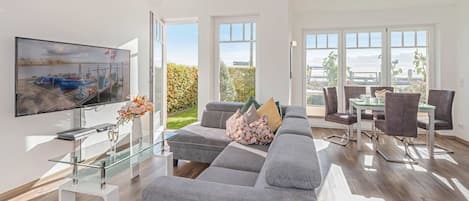 A modern living room with a grey sofa, a glass coffee table, a flat-screen TV, and a dining area with a table and chairs.