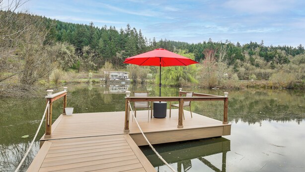 A wooden dock with a red umbrella, chairs, and a table by a calm lake with forested hills in the background. - Private Pond & Dock! Cozy Cabin in Southern Oregon (Oakland)