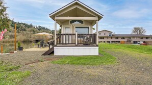 A small, single-story building with a porch, a covered outdoor seating area, and a view of a lake. - Private Pond & Dock! Cozy Cabin in Southern Oregon (Oakland)