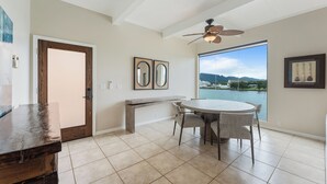 A spacious kitchen with a large dining table, a ceiling fan, and a view of a lake through a large window. - Oceanview Kauai Retreat | Balcony & Beach Access (Lihue)