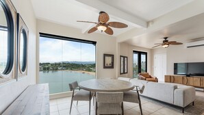 A modern living room with a large window, a round dining table, a sofa, and two ceiling fans. - Oceanview Kauai Retreat | Balcony & Beach Access (Lihue)