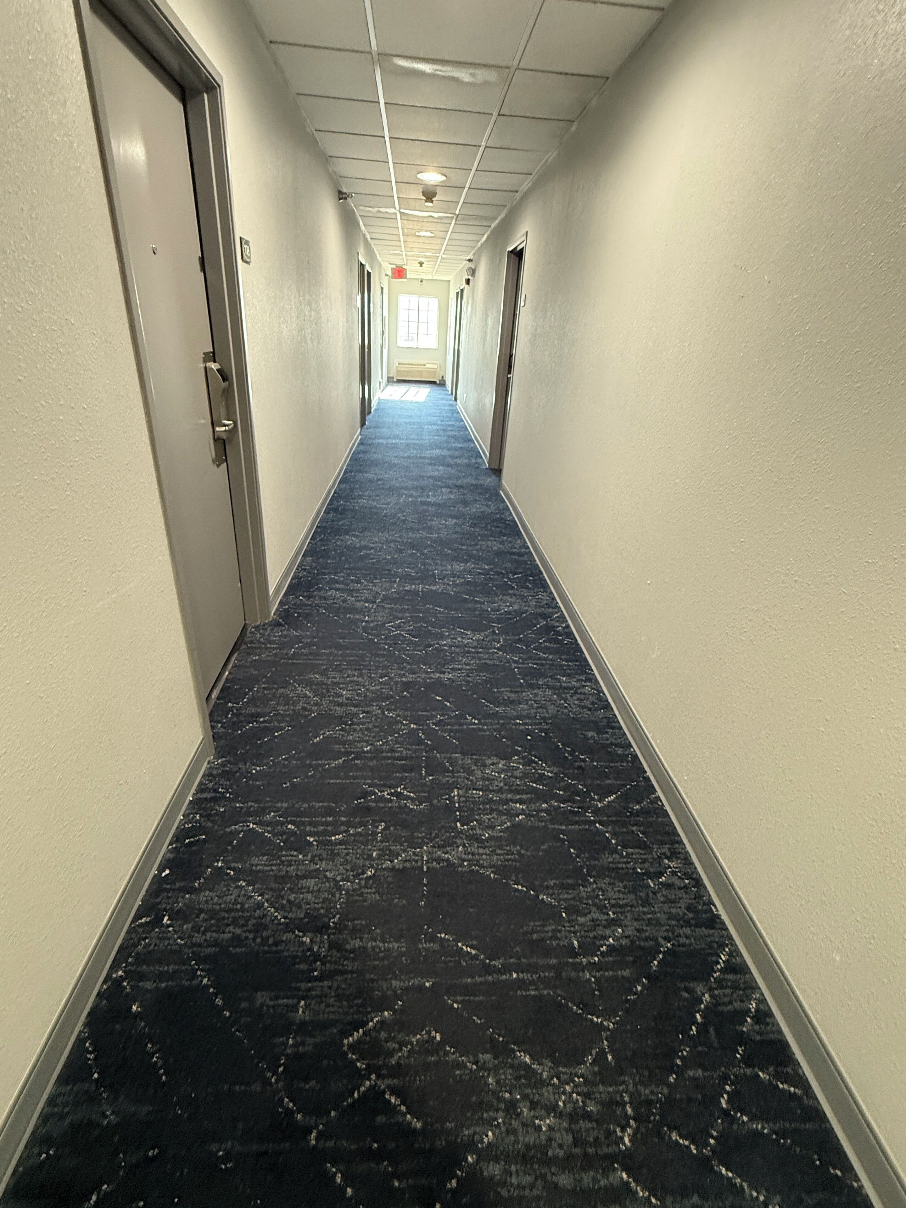 Un pasillo largo y estrecho con alfombra estampada, paredes blancas y puertas a ambos lados.