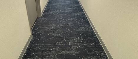 Un pasillo largo y estrecho con alfombra estampada, paredes blancas y puertas a ambos lados.
