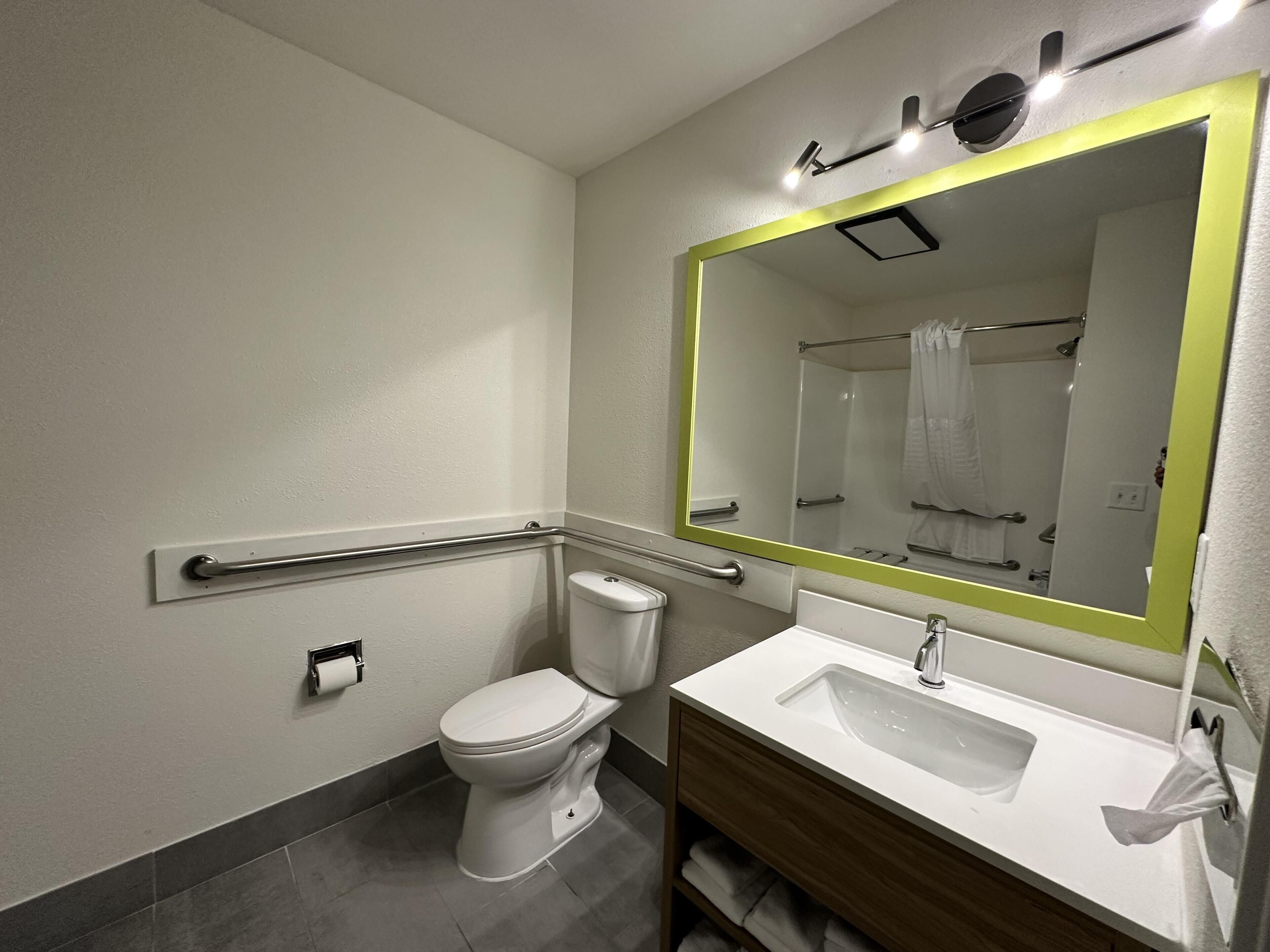 A modern bathroom with a toilet, sink, mirror, and handrail.